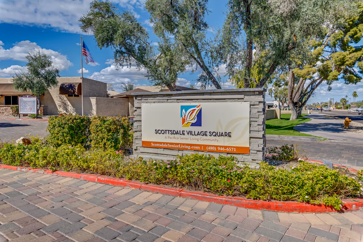 Outdoor view of the entrance sign for Scottsdale Village Square, a Pacifica Senior Living Community, surrounded by greenery and trees with a paved driveway and an American flag in the background under a blue sky with clouds.
