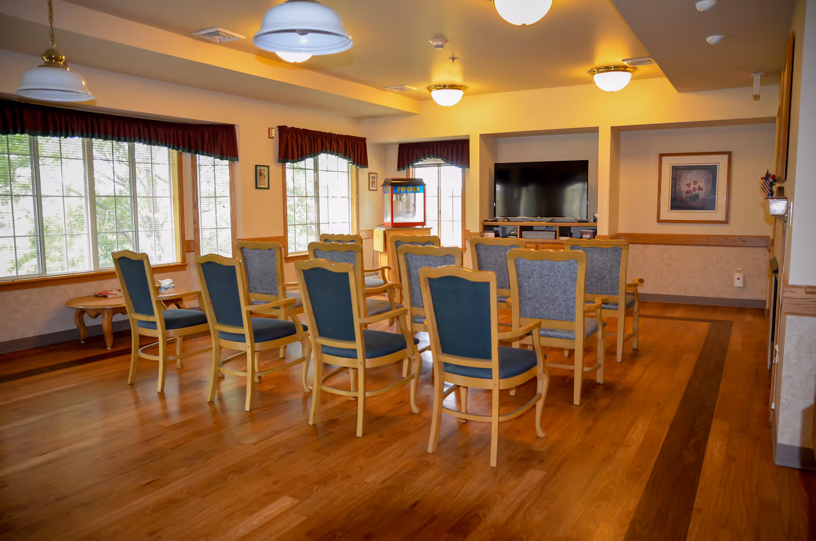 A bright common room with rows of wooden chairs facing a television and large windows.