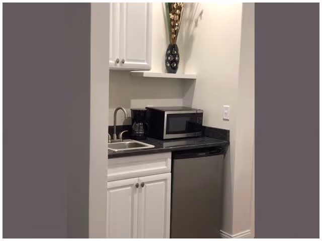 A small kitchenette area with white cabinets, a stainless steel sink, a coffee maker, a microwave, and a stainless steel mini refrigerator. A decorative vase with dried plants is placed on a shelf above the microwave.