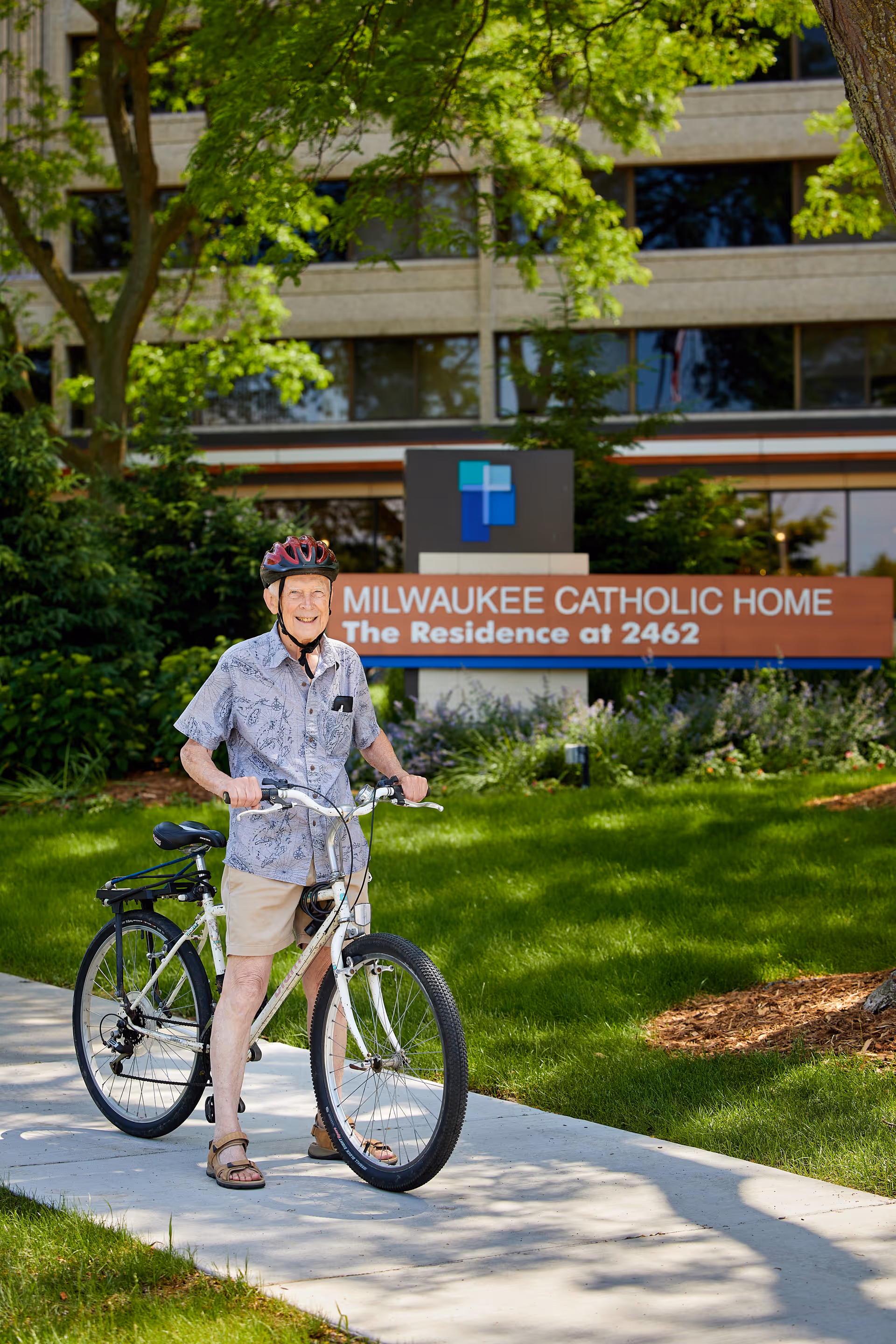 An elderly man wearing a helmet, a short-sleeve button-up shirt, shorts, and sandals is standing with a bicycle on a sidewalk in front of a building. Behind him is a sign that reads 'Milwaukee Catholic Home The Residence at 2462' with greenery and trees surrounding the area.