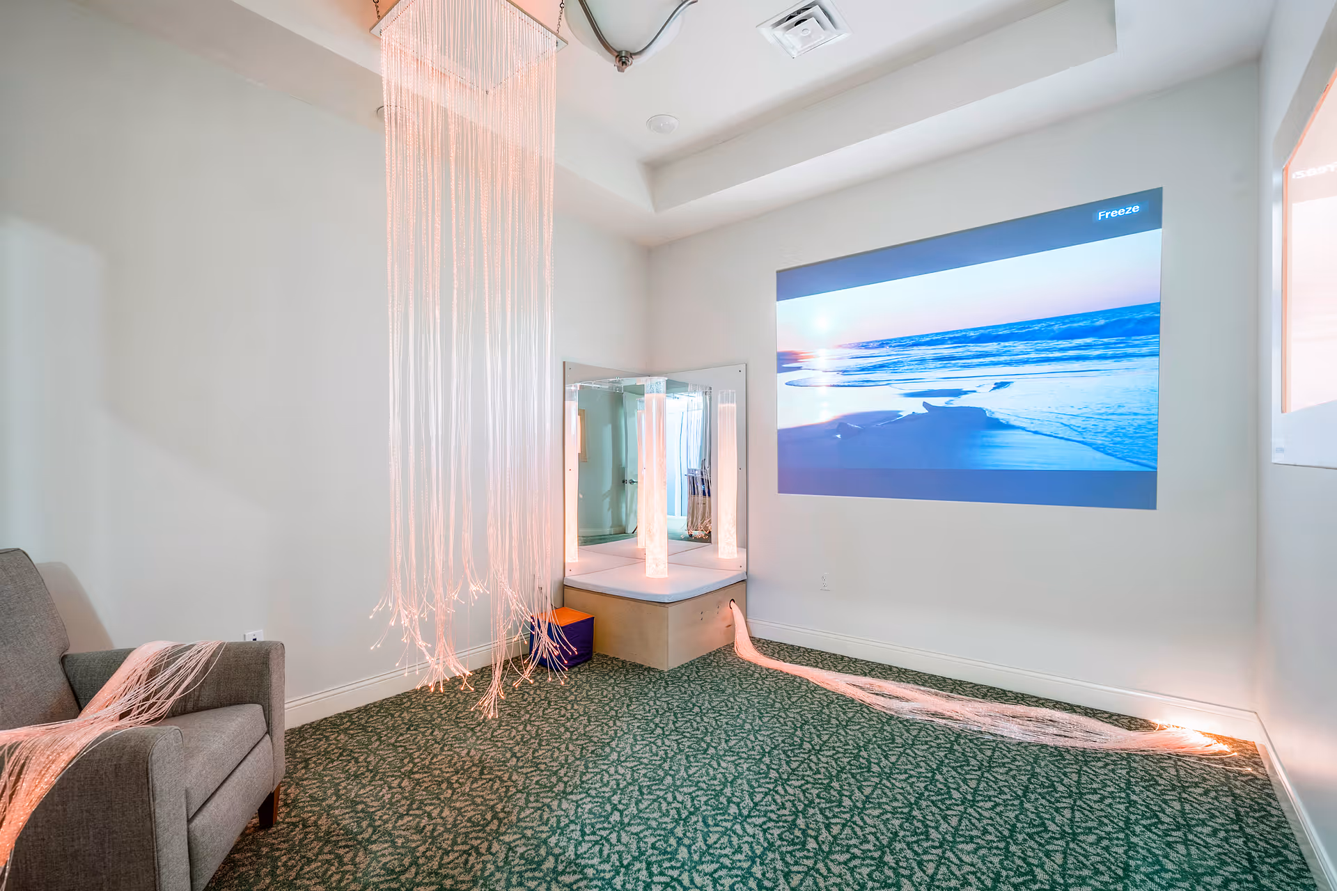 A calm interior sensory room with hanging fiber-optic lights, a beach projection on the wall, an armchair and patterned green carpet.