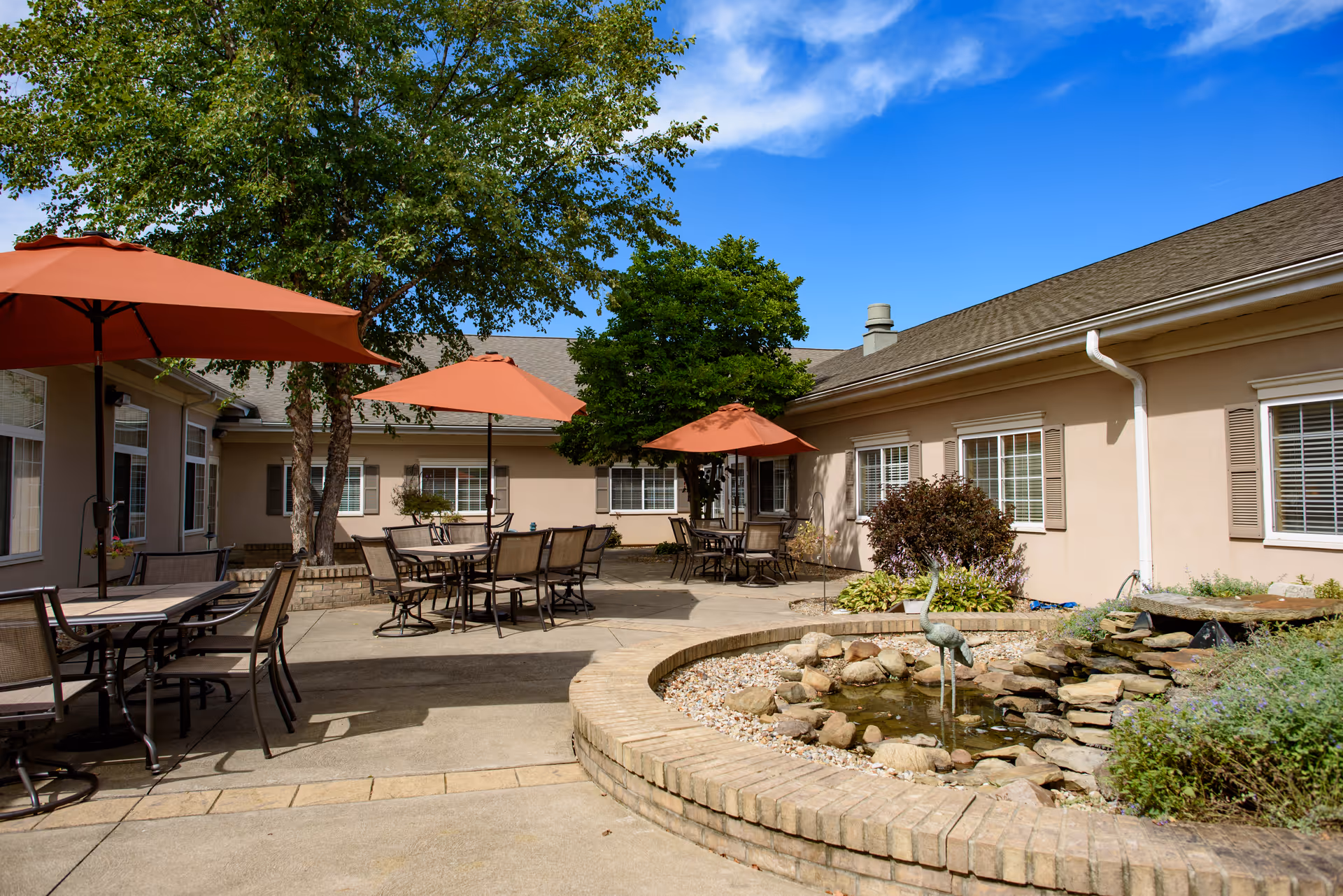 Outdoor courtyard area with multiple tables and chairs under orange umbrellas. There are trees and shrubs around, and a small stone water feature with a bird sculpture in the foreground. The courtyard is surrounded by a beige building with multiple windows and a clear blue sky above.