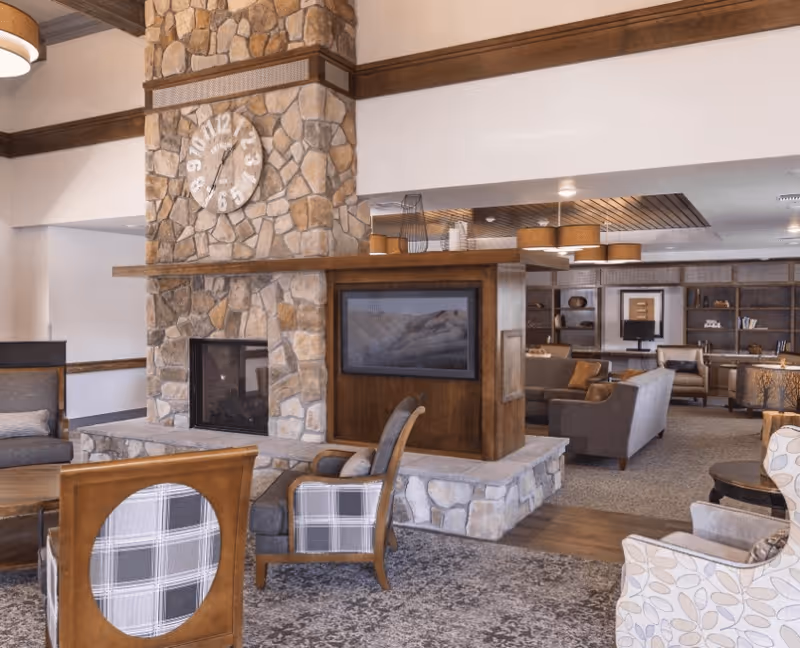A cozy living room area featuring a large stone fireplace with a clock mounted above it. The fireplace has a wooden mantel and a built-in television on one side. The room is furnished with various upholstered chairs and sofas in neutral tones, arranged on a patterned carpet. In the background, there are bookshelves and additional seating, with warm lighting from ceiling fixtures.