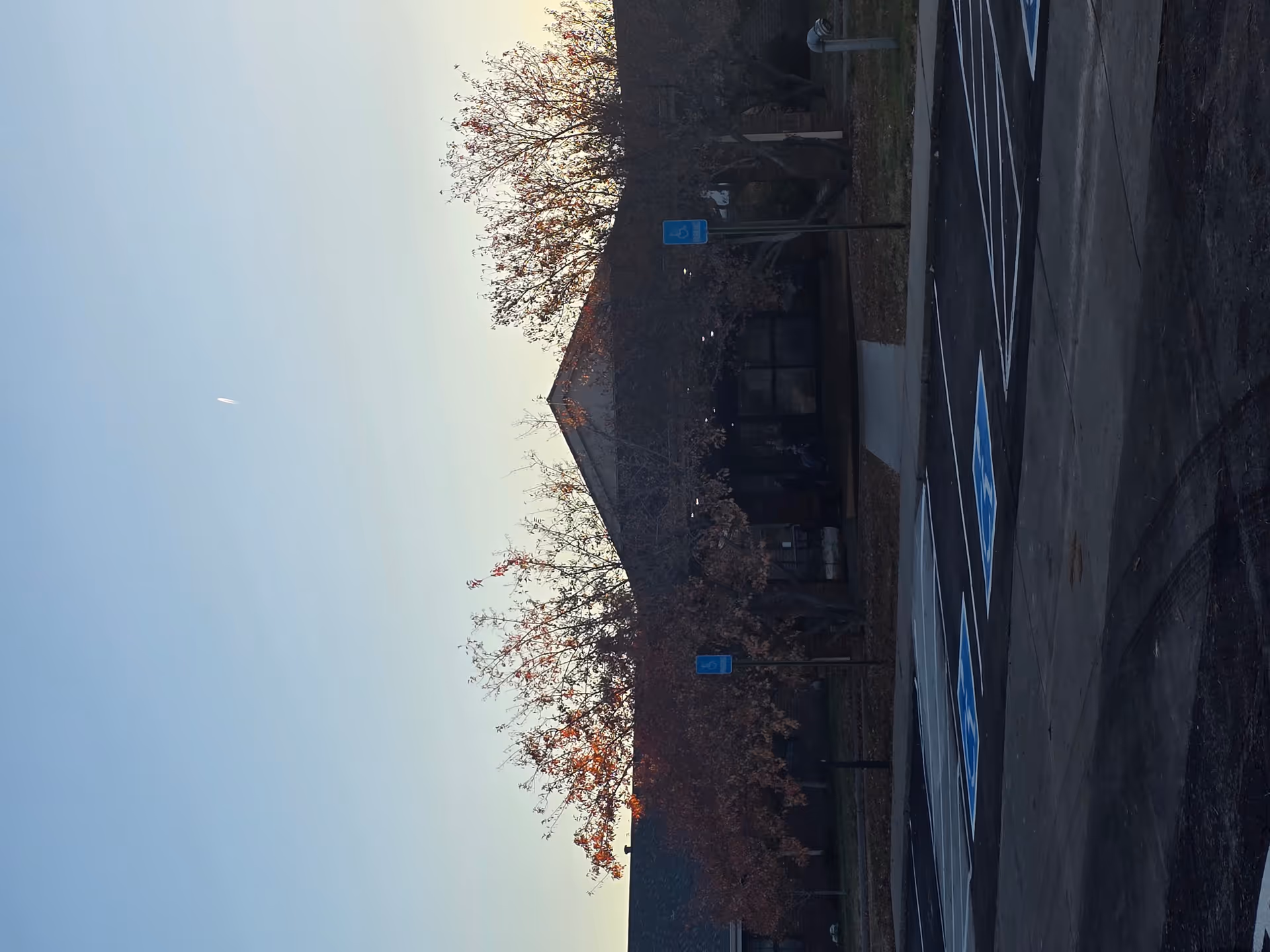 Exterior view of a building partially obscured by leafless trees with a parking lot in the foreground. The parking lot has several handicap parking spaces marked with blue signs and symbols on the ground. The sky is clear and light blue.