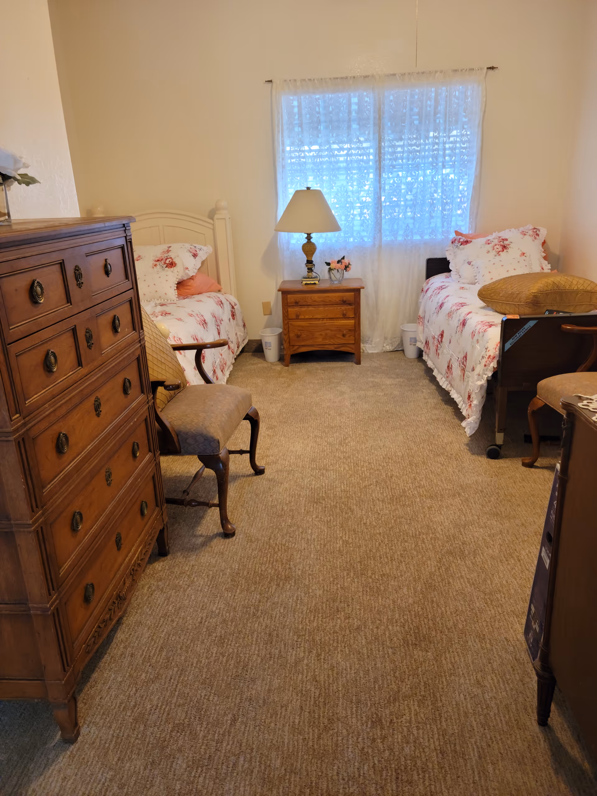 Carpeted bedroom with two floral twin beds, a nightstand with a lamp between them, wooden dressers and chairs.