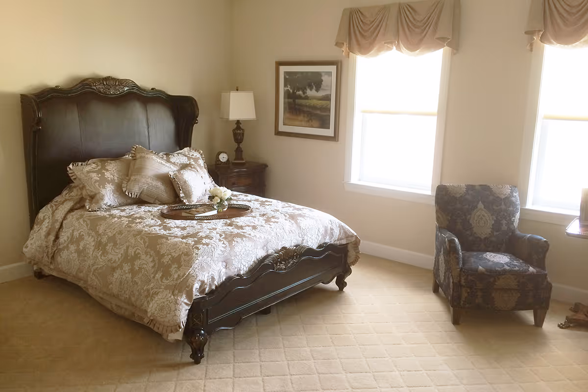 A bedroom with a dark wooden bed frame and a beige patterned bedspread with matching pillows. There is a wooden nightstand with a lamp and a clock next to the bed. Two windows with beige valances let in natural light. A framed landscape painting hangs on the wall between the windows. A patterned armchair is placed near the windows on a beige carpeted floor.