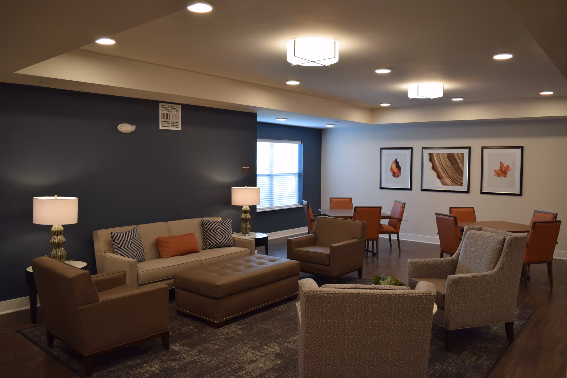A cozy senior living facility common area with a beige sofa, two brown armchairs, a large brown ottoman, and two side tables with lamps. In the background, there are two tables with orange chairs and three framed pictures of leaves and wood patterns on the wall. The room has recessed lighting and a window with blinds.