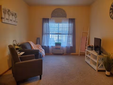 A cozy living room with beige walls and carpeted floor. The room features a gray armchair and a gray sofa with a pink throw blanket. A window with sheer white and purple curtains lets in natural light. There is a white TV stand with a flat-screen TV, a small potted plant, and a decorative ladder with a pink blanket. A wall clock and framed artwork are visible on the walls.