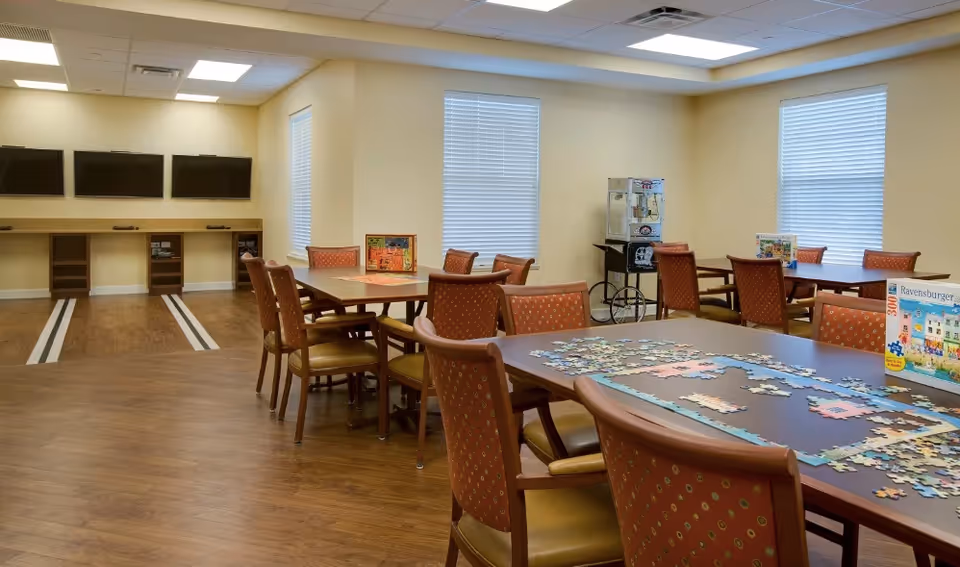 A well-lit activity room with several tables and chairs arranged for group activities. On the tables, there are puzzle pieces and puzzle boxes. The room has wooden flooring, beige walls, and three large windows with blinds. On one wall, there are three mounted flat-screen TVs above a wooden shelf unit. A popcorn machine on a cart is placed near one of the windows.