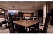 Interior view of a dining area in a senior living facility with a square wooden table surrounded by dark wooden chairs. A small floral centerpiece is placed on the table. The room has carpeted flooring, recessed lighting in the ceiling, and a warm, inviting ambiance.