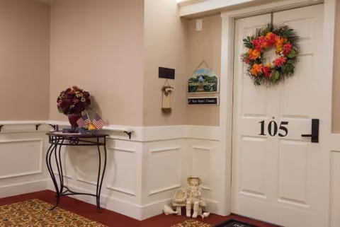 Interior hallway corner of an assisted living facility with a white door numbered 105 decorated with a colorful flower wreath. A small black metal table with a flower arrangement and two small American flags is placed against the wall. Below the table, there are two small decorative dolls sitting on the floor. The walls are beige with white wainscoting and a patterned carpet on the floor.