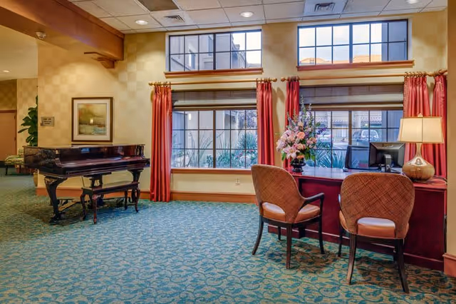 Interior view of a senior living facility common area with a grand piano on the left, a desk with two chairs on the right, a lamp, and a flower arrangement on the desk. Large windows with red curtains let in natural light, and the room has patterned carpet and beige walls.