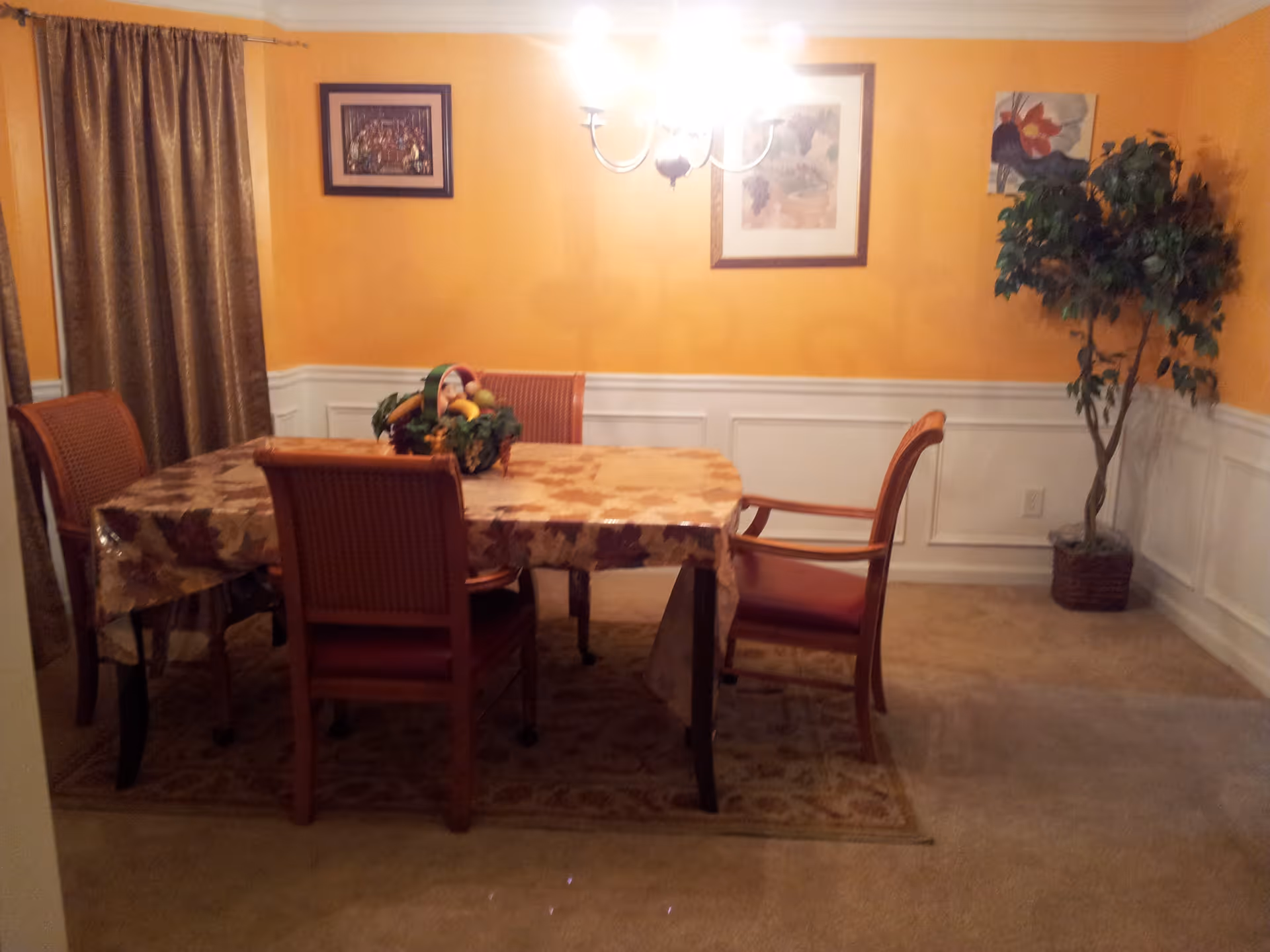 A dining room with a rectangular table covered with a patterned tablecloth and four wooden chairs with red cushions. A fruit basket centerpiece is on the table. The walls are painted yellow with white wainscoting, and there are three framed pictures hanging on the walls. A potted artificial tree stands in the corner, and a chandelier with five lights hangs above the table. Brown curtains cover a window on the left side.