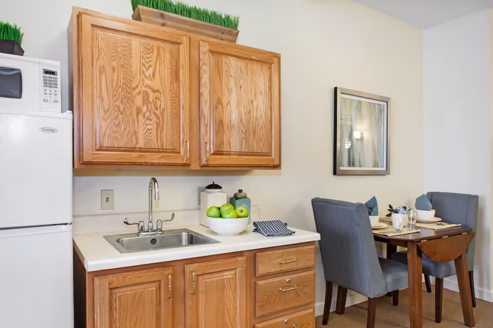 A small kitchen area with wooden cabinets, a white countertop with a sink, a bowl of green apples, and a folded dish towel. To the left is a white refrigerator with a microwave on top. To the right, there is a small dining table set for two with gray upholstered chairs and a framed abstract painting on the wall.