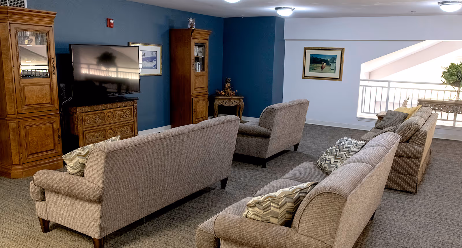 Community lounge with upholstered sofas and armchairs facing a TV and wooden cabinets in a carpeted room.