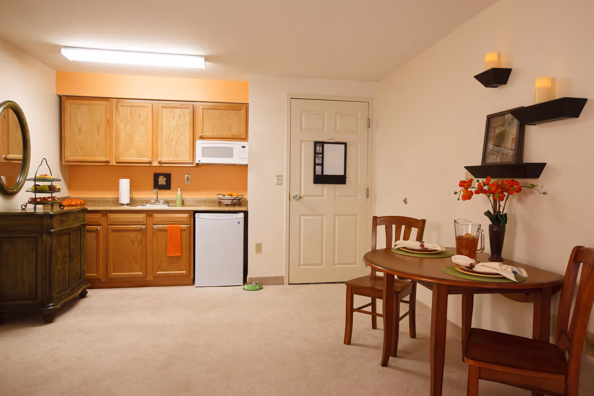 A small kitchen and dining area in a senior living facility. The kitchen features wooden cabinets, a microwave, a mini refrigerator, a sink, and a countertop with a paper towel holder and some fruit. To the right, there is a round wooden dining table set for two with plates, napkins, and a pitcher of iced tea. The wall above the table has two floating shelves with decorative candles and a framed picture. A door is visible in the background, and the room has beige walls and carpeted flooring.
