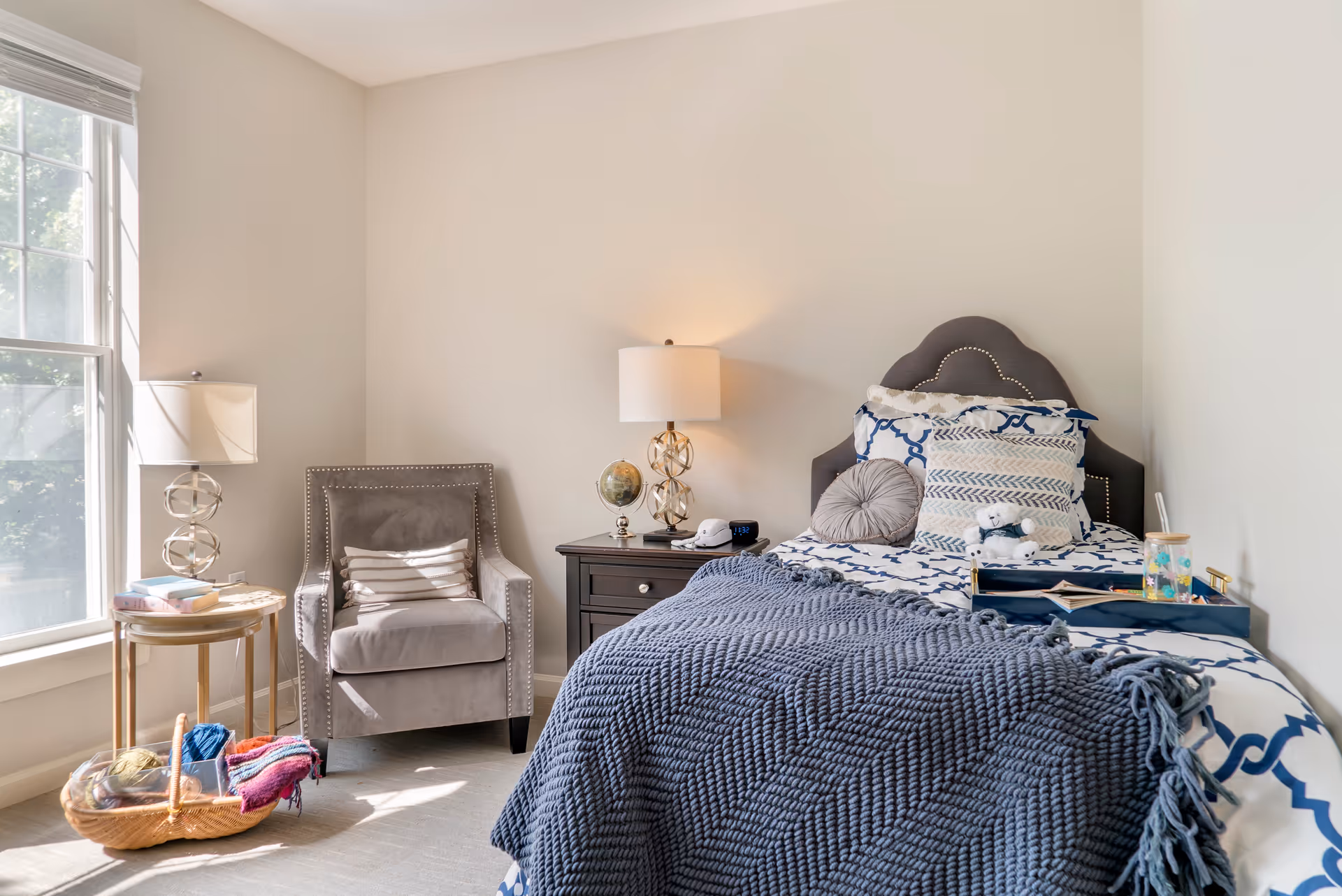 A cozy bedroom with a single bed featuring a dark gray headboard, blue and white patterned bedding, and multiple pillows including a round gray one and a small teddy bear. A dark wooden nightstand beside the bed holds a decorative globe, a lamp with a white shade, and a digital clock. Next to the nightstand is a gray upholstered armchair with a striped pillow. A small round side table with a lamp and some books is positioned near a large window letting in natural light. A basket with yarn and knitting supplies is on the floor near the chair.