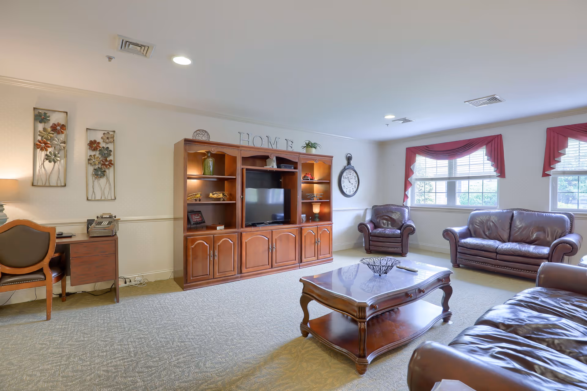 A spacious living room with beige carpet and cream-colored walls. The room features a wooden entertainment center with a flat-screen TV, decorative items, and the word 'HOME' displayed on top. There are three leather armchairs and a matching leather sofa arranged around a wooden coffee table. Two windows with red valances allow natural light to brighten the room. On the left side, there is a small wooden desk with a chair and a lamp, and two floral wall decorations above it. A large wall clock is mounted near the windows.