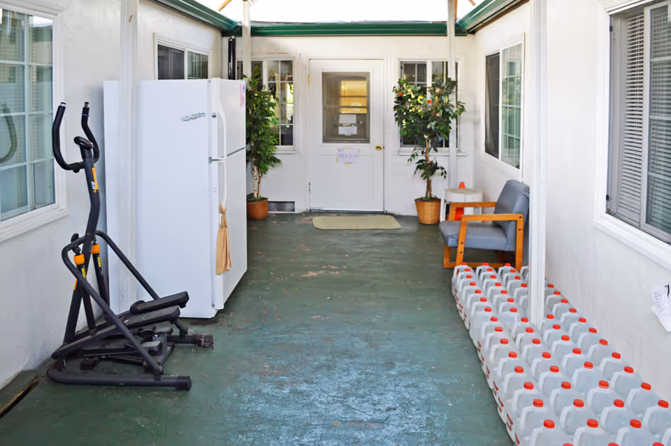 Enclosed corridor with a refrigerator and exercise machine on the left, a chair and potted plants near a door at the far end, and a row of plastic water jugs along the right side.