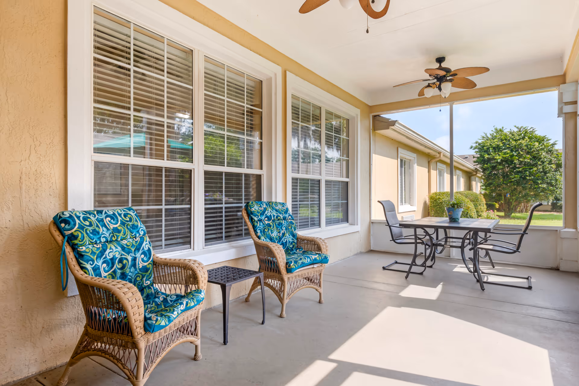A covered outdoor patio area with two wicker chairs featuring blue patterned cushions, a small black side table between them, and a black metal table with two chairs. The patio has ceiling fans with light fixtures and overlooks a green lawn with bushes and a tree.