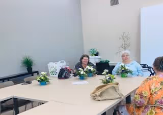 Three elderly women sitting around a large beige table in a bright room with white walls. The table has small flower arrangements and personal items like bags and a laundry basket. There is a small plant on a bench against the wall.