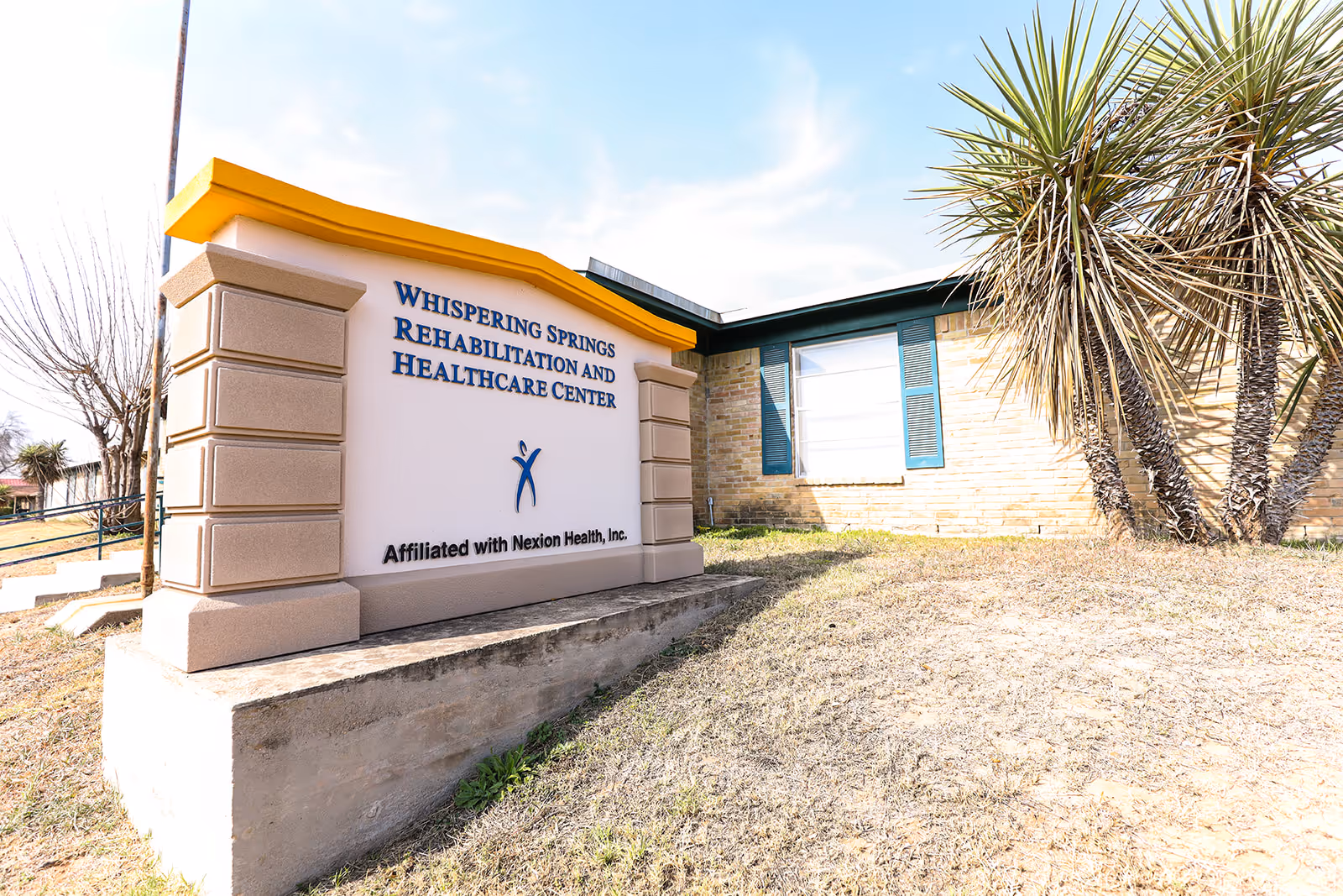 Outdoor sign reading "Whispering Springs Rehabilitation and Healthcare Center" in front of a low brick building with landscaping.