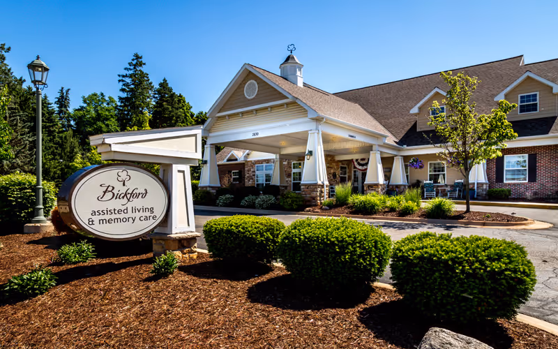 Exterior view of Bickford Of Okemos assisted living and memory care facility with a large covered entrance, manicured bushes, and a sign displaying the facility's name. The building has a brick and siding facade with a peaked roof and a clear blue sky in the background.