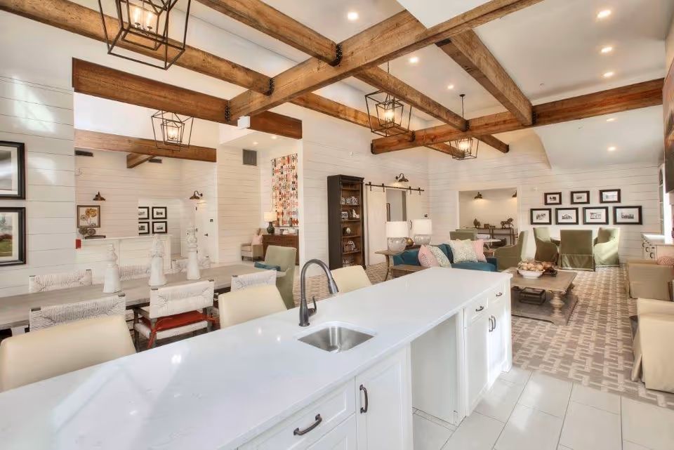 Bright and spacious interior of a senior living facility featuring a modern kitchen island with a sink in the foreground, a dining area with a large table and chairs to the left, and a comfortable living room with sofas, armchairs, and framed pictures on the walls. The ceiling has exposed wooden beams and pendant lights, and the floor is tiled with a patterned rug in the living area.