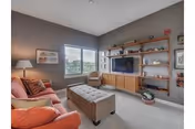 A cozy living room with a coral sofa adorned with pillows, a tufted ottoman in the center, a wooden chair near two large windows, and a wall-mounted flat-screen TV surrounded by wooden shelves holding decorative items.
