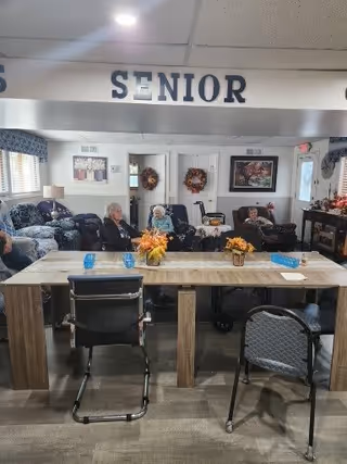 A senior living facility common area with several elderly residents seated in armchairs along the back wall. In the foreground, there is a long wooden table with chairs around it, decorated with small flower arrangements and blue drinking glasses. The room has a cozy atmosphere with framed artwork and wreaths on the walls, and a fireplace on the right side.