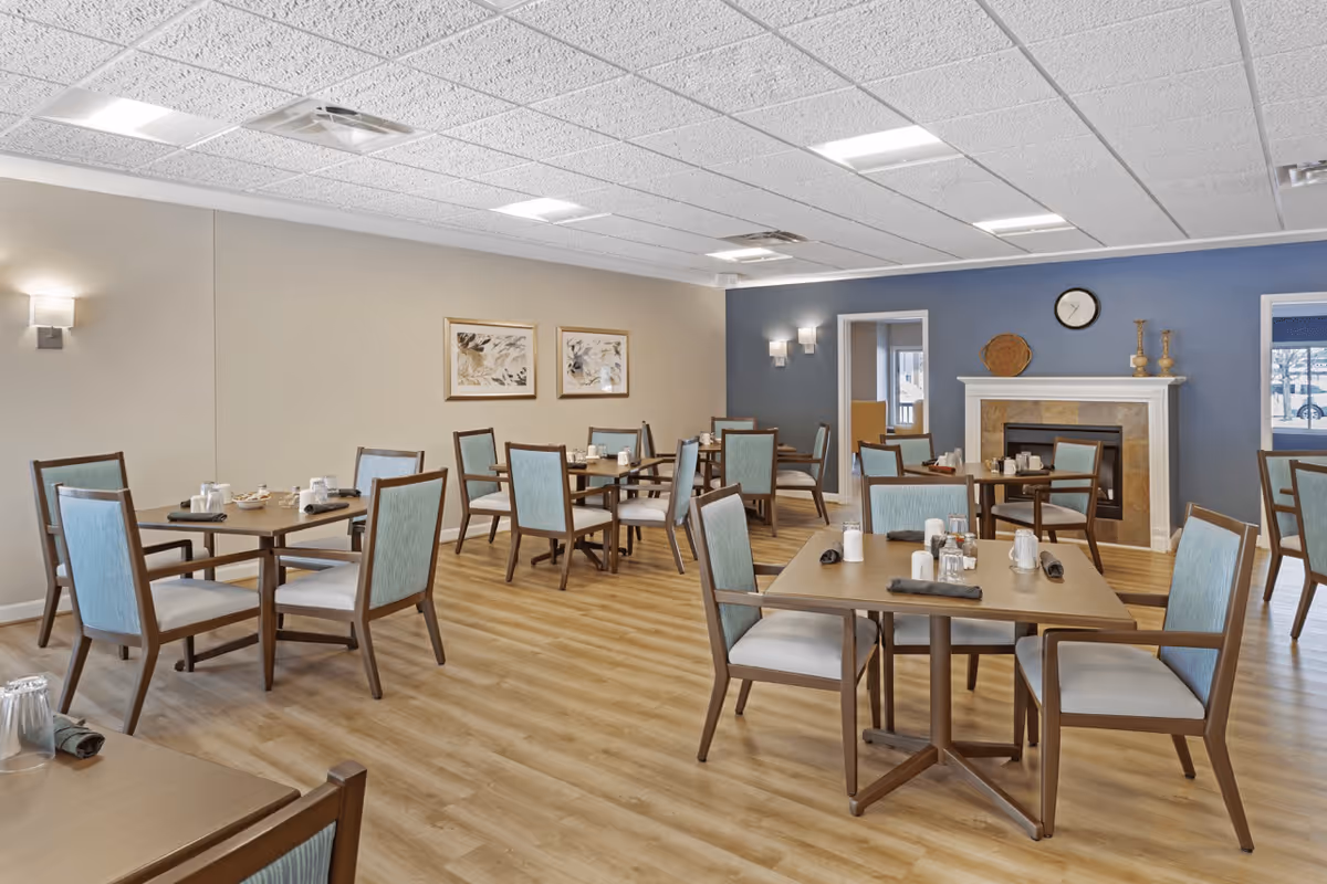 A senior living facility dining room with multiple square tables, each surrounded by four chairs with light blue upholstery. The room has light wood flooring, beige and blue walls, framed artwork, a fireplace with decorative items on the mantel, and ceiling lights.
