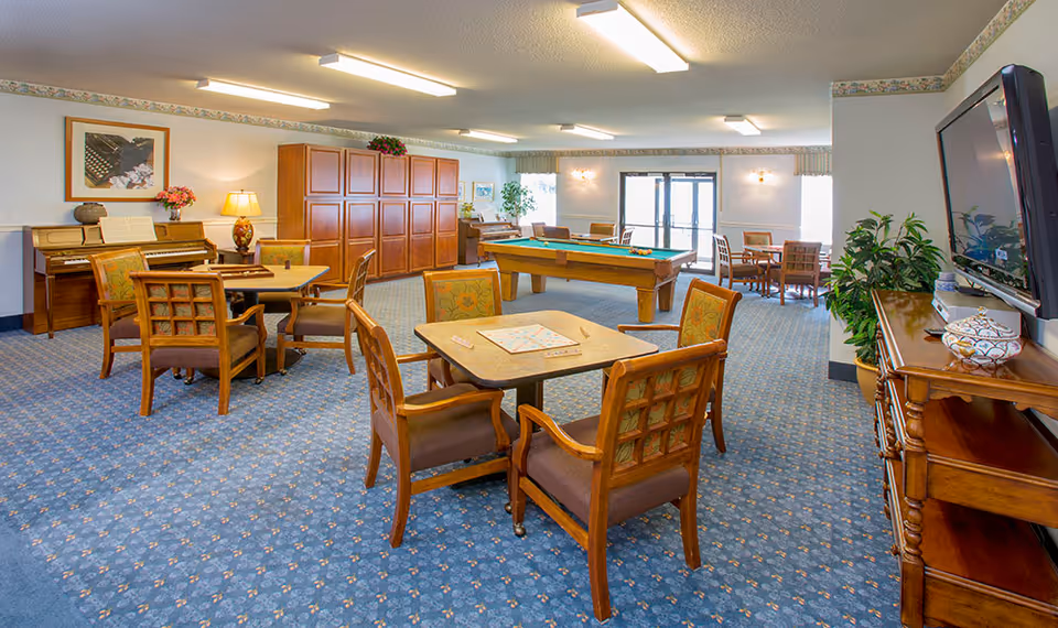 Spacious senior community activity room with tables and chairs, a pool table, piano, and TV.