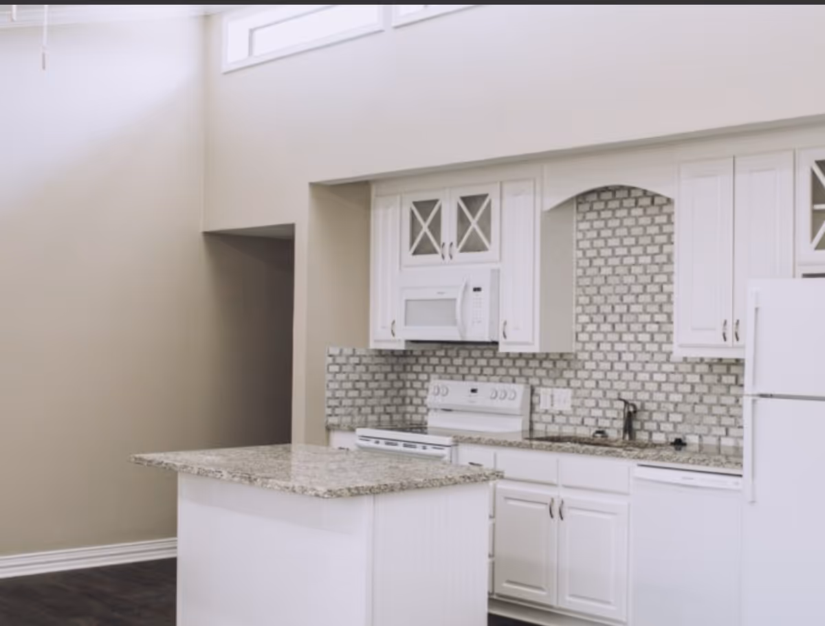 A modern kitchen with white cabinetry, a white refrigerator, a white microwave above a white stove, a dishwasher, and a kitchen island with a granite countertop. The backsplash features a white and gray brick pattern, and the walls are painted beige with a high window letting in natural light.