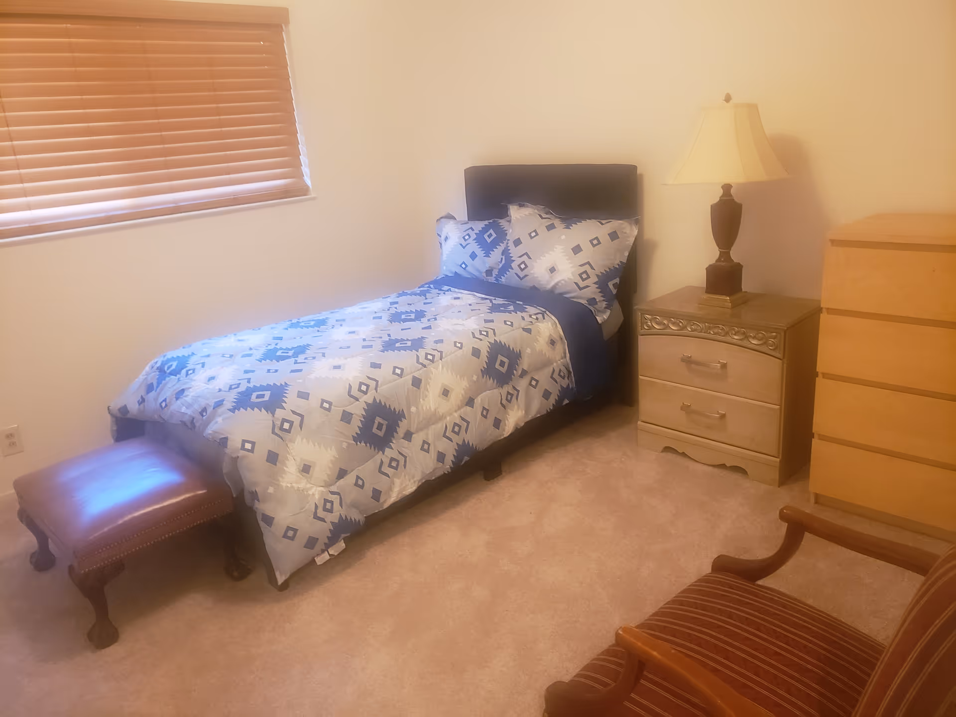 Furnished single bedroom with a twin bed with patterned bedding, a nightstand and lamp, dresser, footstool and chair.