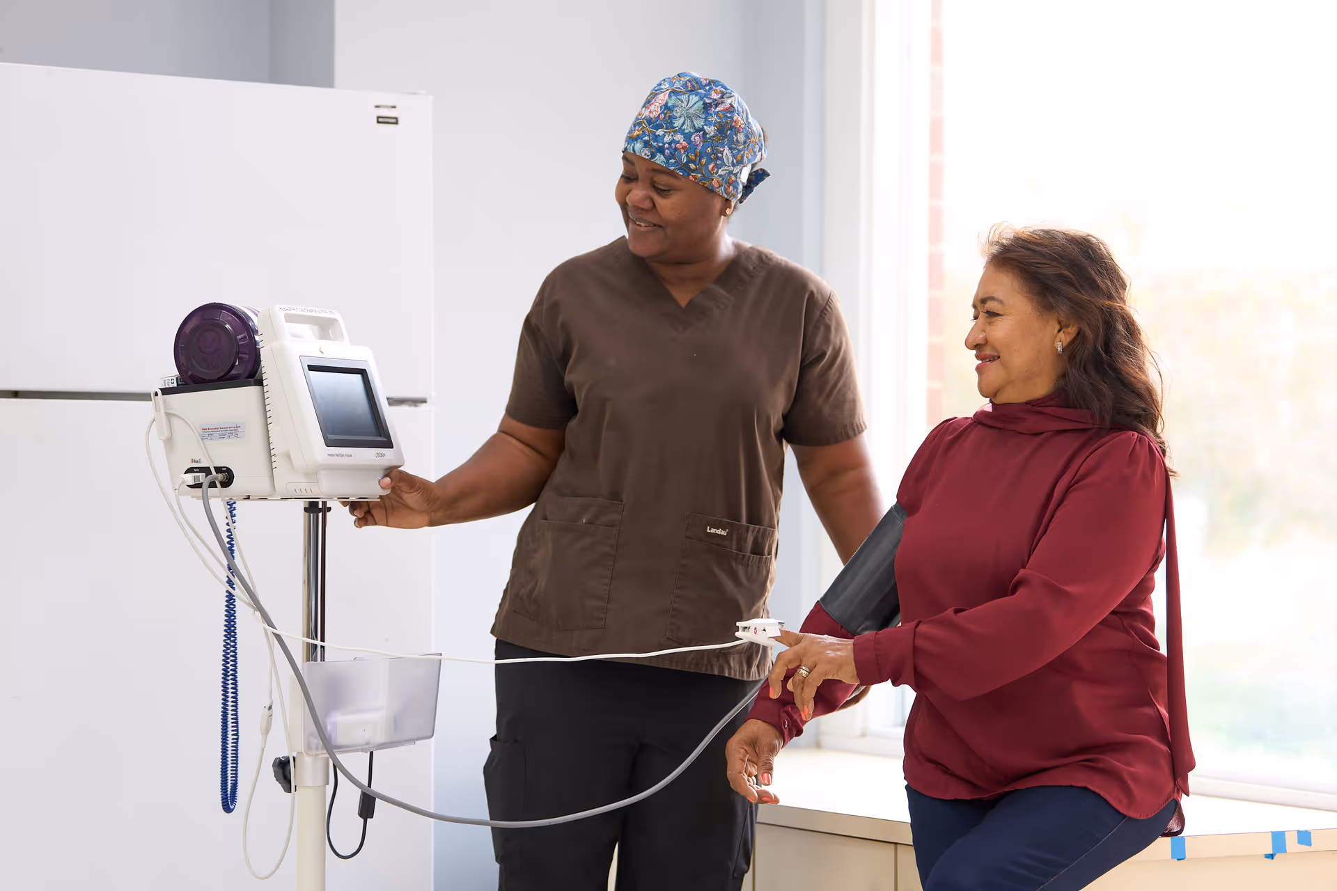 A healthcare worker in brown scrubs and a patterned head covering is taking the blood pressure of a seated woman wearing a maroon blouse. They are indoors near a large window with natural light.