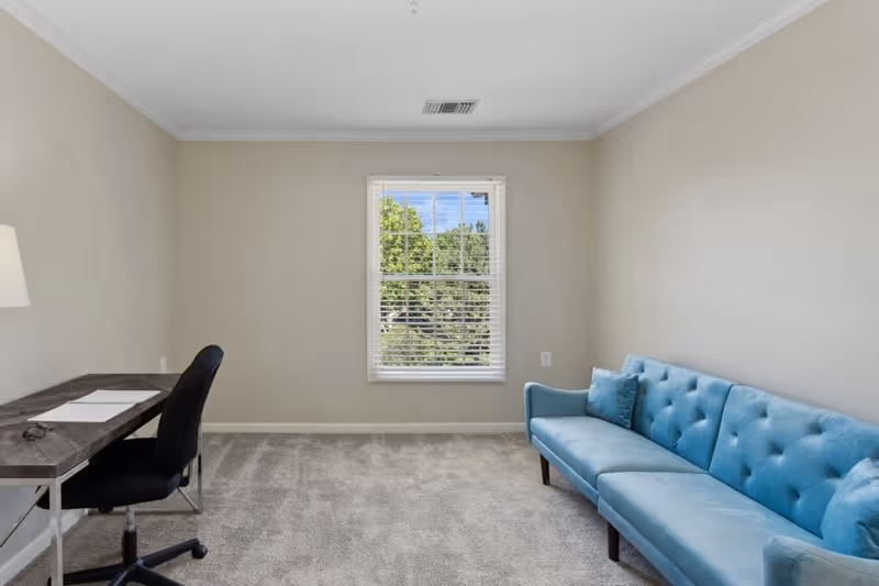 A small room with beige walls and carpeted floor featuring a blue tufted sofa on the right side and a dark wooden desk with a black office chair on the left side. A window with white blinds is centered on the back wall, showing green trees outside.