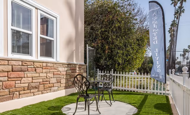 Small outdoor patio with a metal table and two chairs on artificial grass beside a building, white picket fence, and a vertical banner.