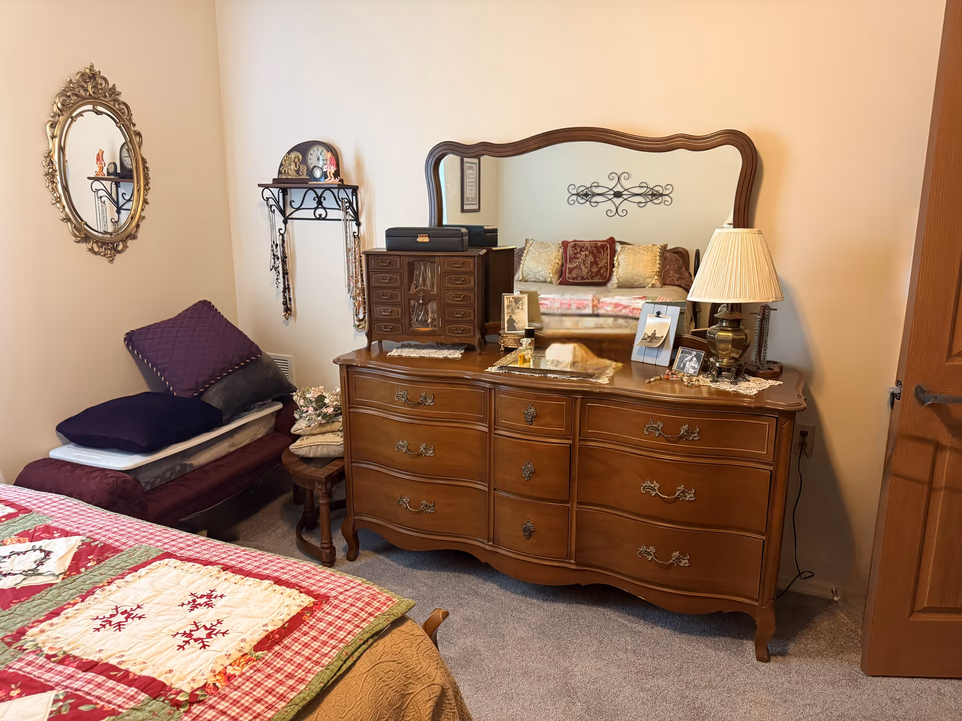 A cozy bedroom corner featuring a wooden dresser with an ornate mirror, a table lamp, framed photos, and decorative items on top. To the left, there is a small bench with folded blankets and pillows, a round wooden side table, and a wall-mounted shelf holding a clock and hanging necklaces. The bed with a patchwork quilt is partially visible in the foreground.