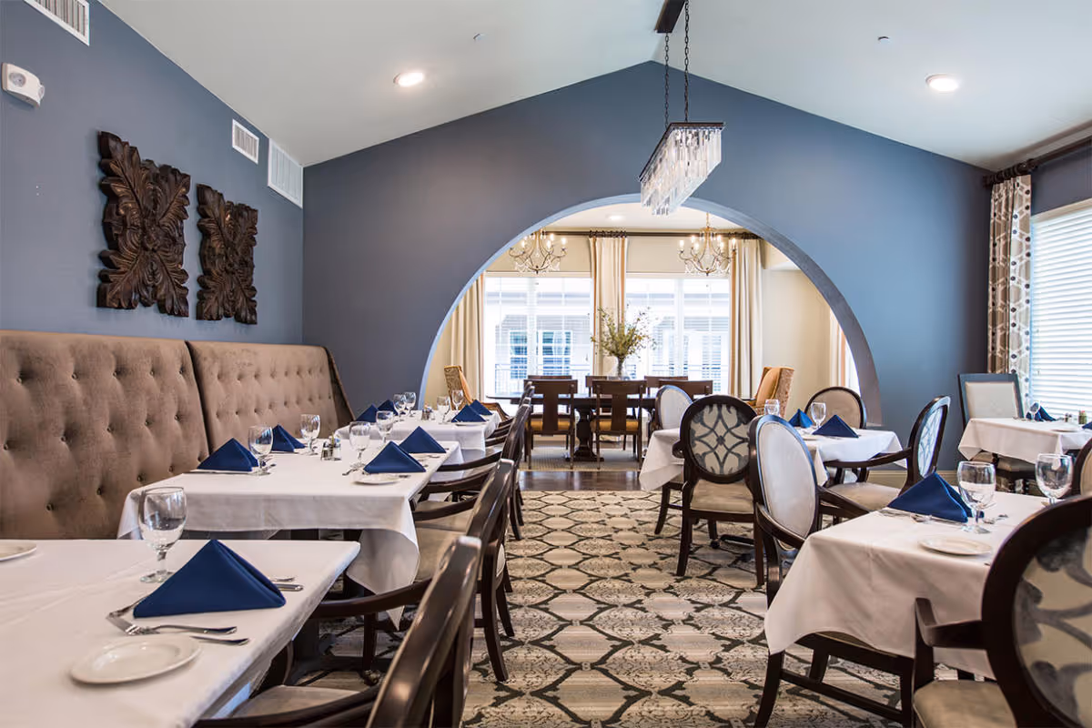Elegant dining room with tables covered in white tablecloths, each set with blue folded napkins, plates, glasses, and silverware. The room features a tufted beige bench along one wall, decorative wall art, a patterned carpet, and a large archway leading to another dining area with chandeliers and large windows with curtains.