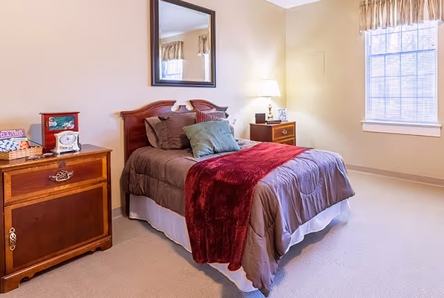 A cozy bedroom with a double bed featuring brown bedding, a red throw blanket, and multiple pillows. There is a wooden nightstand with a lamp and picture frame on the right side of the bed, and a wooden dresser with a clock and basket on the left. A large window with curtains lets in natural light, and a mirror hangs on the wall above the bed.