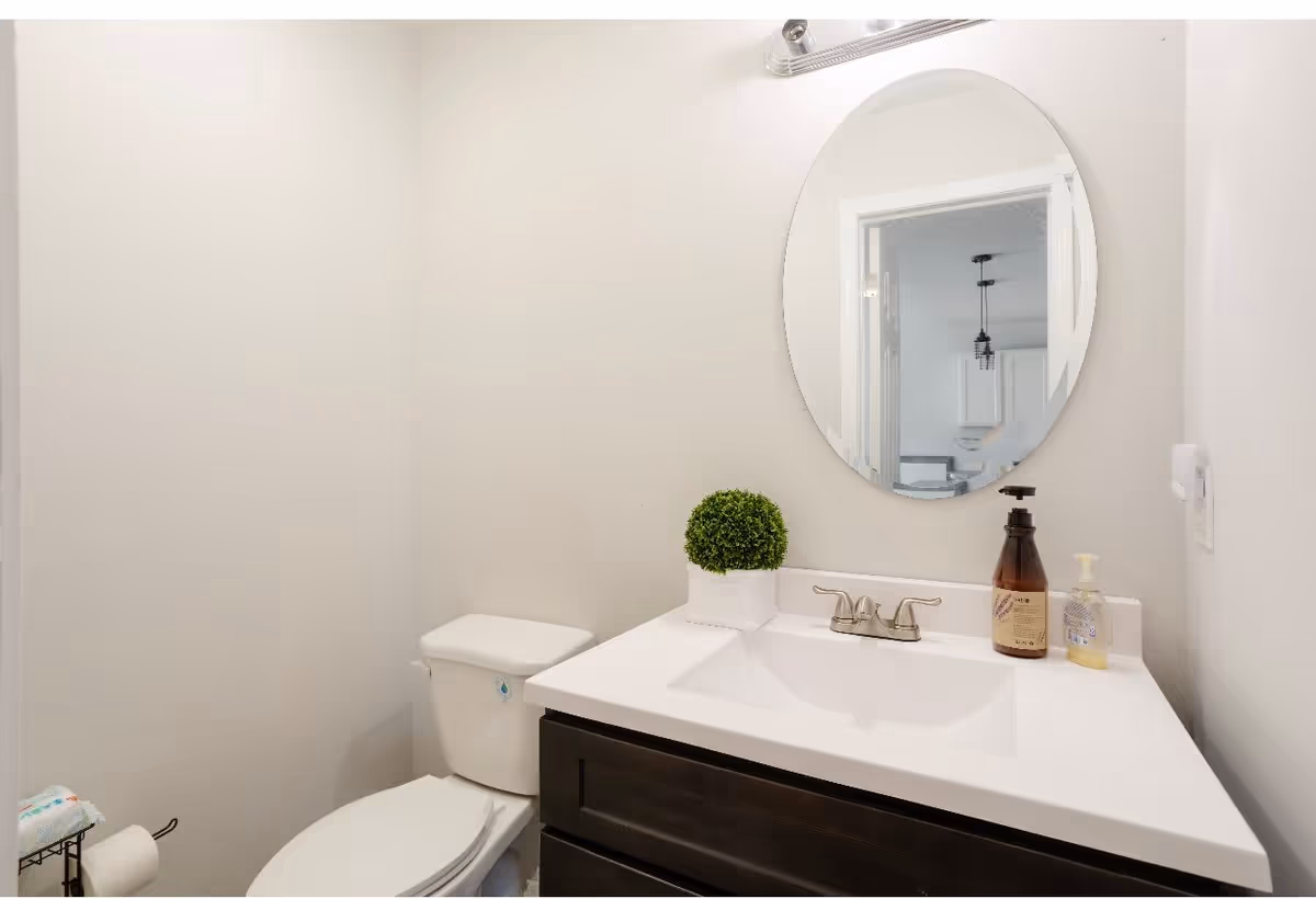 A small bathroom with a white toilet and a dark wood vanity with a white countertop. On the countertop are two soap dispensers and a small green potted plant. Above the vanity is a round mirror reflecting part of an adjacent room with white cabinets and a hanging light fixture. The walls are painted light beige.