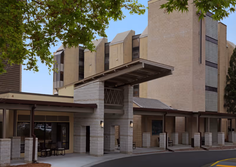 Exterior view of a multi-story assisted living facility building with a covered entrance and seating area. The building has beige brick walls and large windows. Green tree branches frame the top left corner of the image under a clear blue sky.