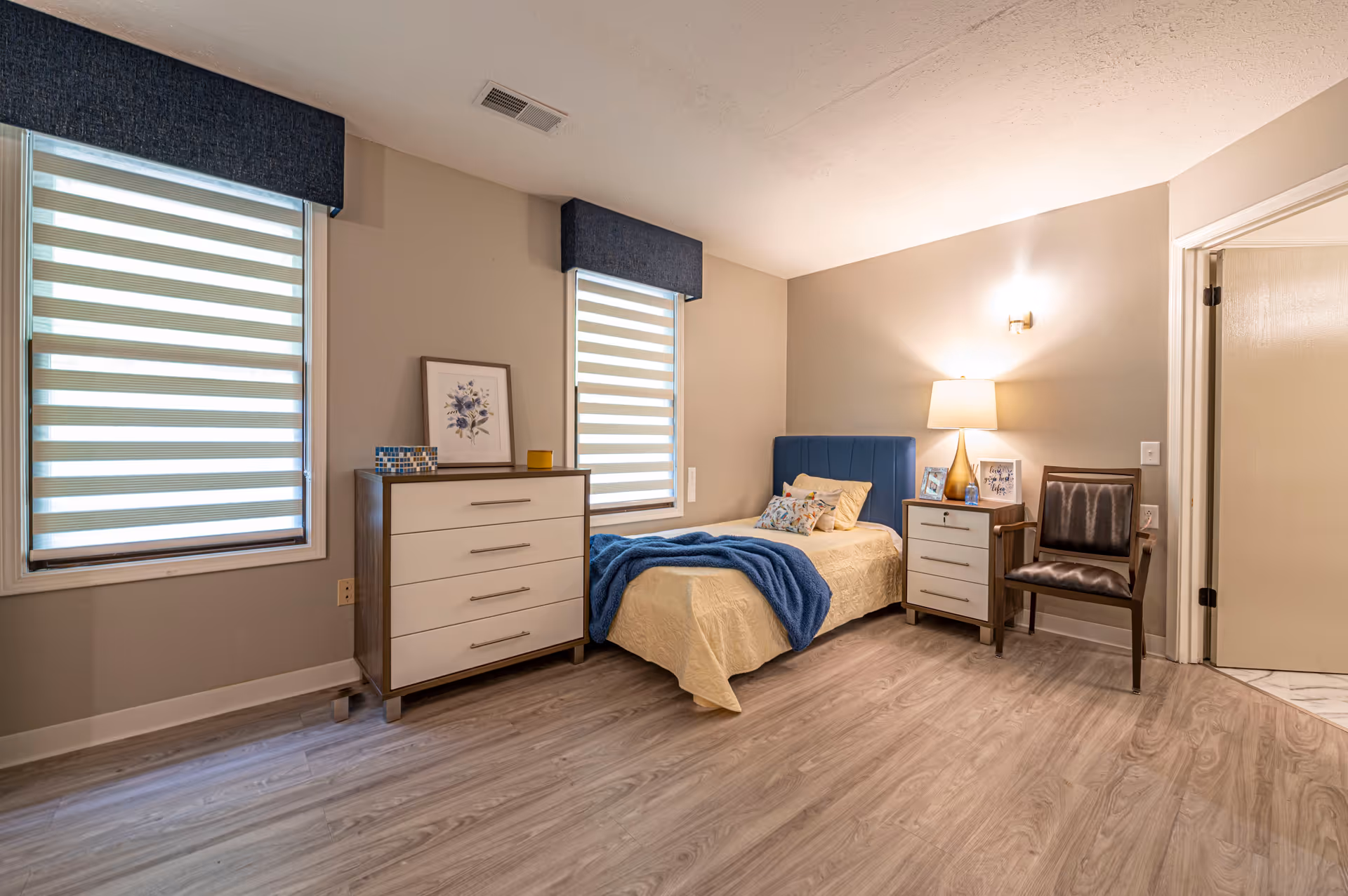 A neatly arranged bedroom with a single bed featuring a blue headboard and beige bedding with a blue throw blanket. There are two windows with striped blinds and dark blue valances. Next to the bed is a nightstand with a lamp, framed photos, and a chair beside it. On the opposite side of the bed is a chest of drawers with decorative items and a framed floral picture on top. The room has light-colored walls and wood flooring.