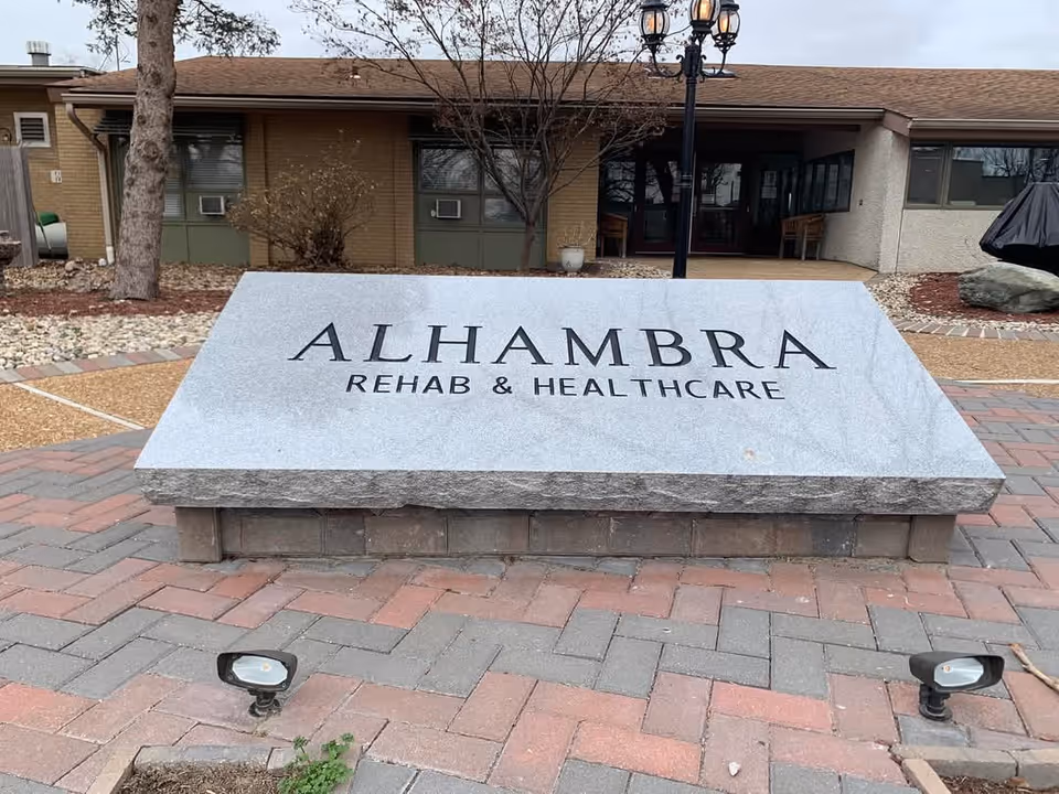 Stone sign with the text 'ALHAMBRA REHAB & HEALTHCARE' in front of a single-story building with windows, a tree, and outdoor lamps.
