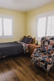 A cozy bedroom with a single bed covered in dark patterned bedding, a wooden nightstand with a lamp, and a colorful patterned armchair. The room has light yellow walls and two windows with white shutters, and a wooden floor.