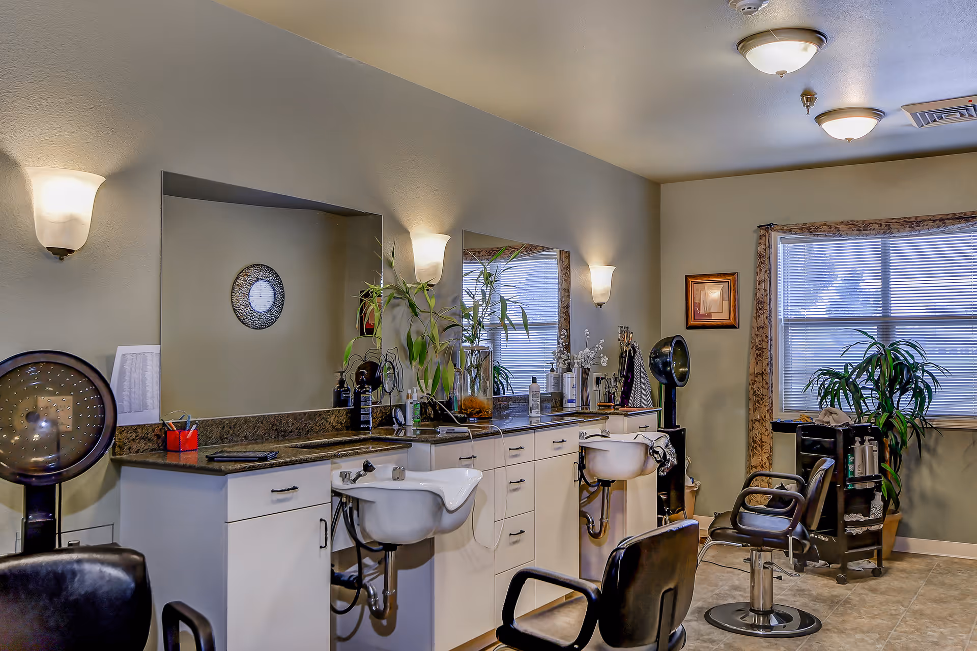 Interior of a hair salon area with two black salon chairs, two white sinks, large mirrors, plants, and various hair care products on the countertop. The room has beige walls, tiled floor, and a window with blinds and curtains.