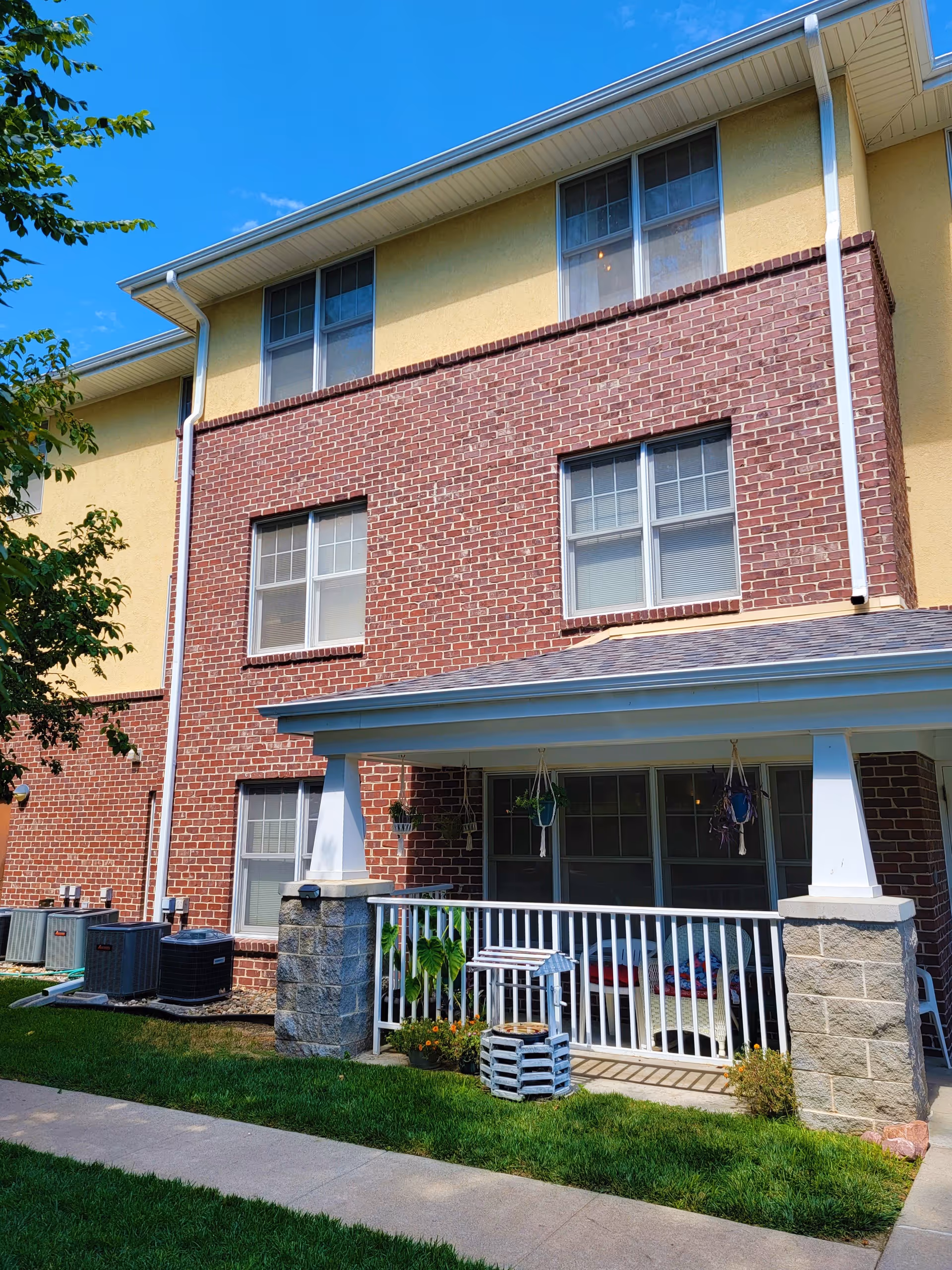 Exterior view of a multi-story senior housing building with a brick and yellow stucco facade. The building features several windows and a covered porch area with white railing, hanging plants, and outdoor seating. There is a well-maintained lawn and a sidewalk in front of the building under a clear blue sky.