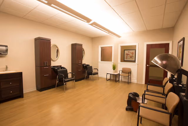 Interior of a hair salon area in a senior living facility with wooden flooring, beige walls, two hair washing stations with mirrors and chairs, a sink with a cabinet, and a row of waiting chairs including one under a hair dryer. There are framed pictures and a small table with a plant near the back wall.