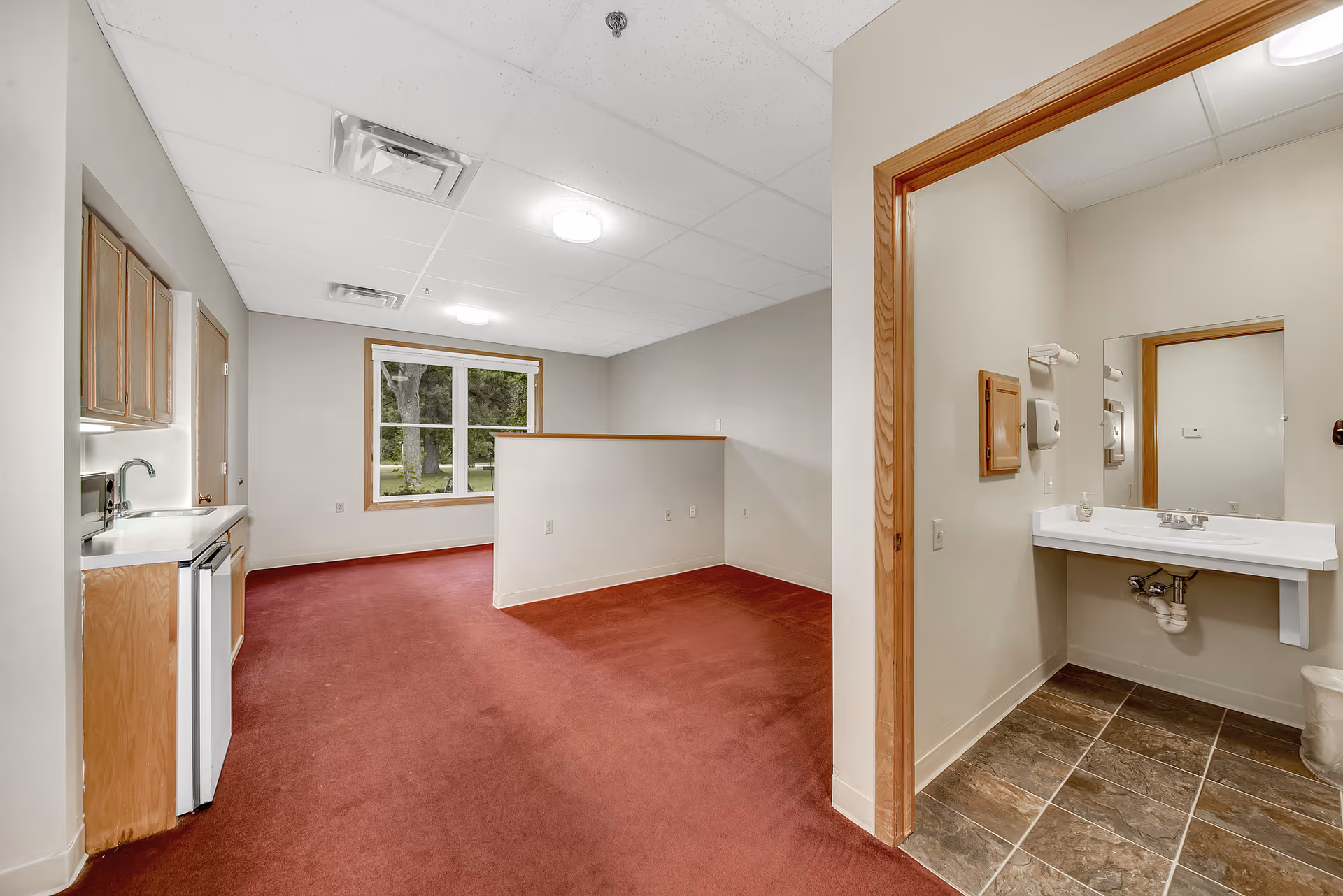 Interior view of a senior living facility room with red carpet flooring, a small kitchenette with wooden cabinets, a mini fridge, and a sink on the left. There is a large window at the back letting in natural light. To the right, there is a bathroom area with a sink, mirror, and tiled floor visible through an open doorway.