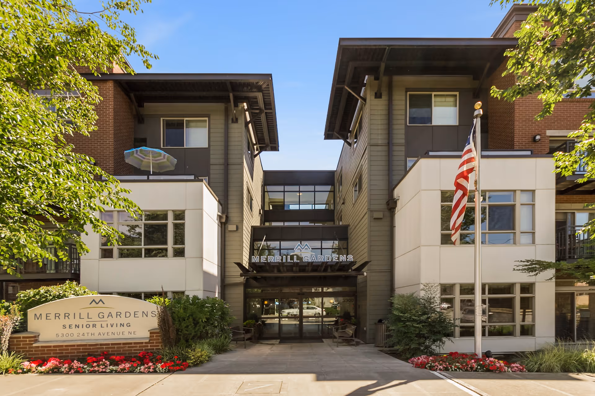 Front exterior view of Merrill Gardens Senior Living facility with a clear blue sky, American flag on a flagpole, and landscaped flower beds. The building has a modern design with large windows and a covered entrance.